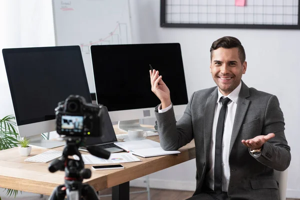 Comerciante sonriente apuntando con la mano durante la transmisión de vídeo en la cámara digital, borrosa primer plano - foto de stock