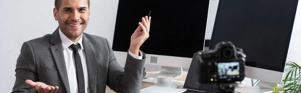 Fröhlicher Händler, der neben Monitoren sitzt und beim Online-Streaming mit der Hand auf Digitalkamera zeigt, verschwommener Vordergrund, Banner — Stockfoto