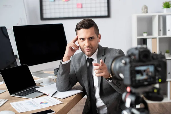 Businessman near laptop and computer monitors pointing with finger at digital camera on blurred foreground — Stock Photo