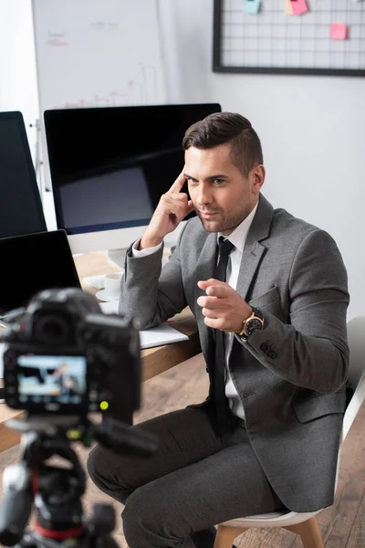 Trader sitting near computer monitors and pointing with finger at digital camera — Stock Photo