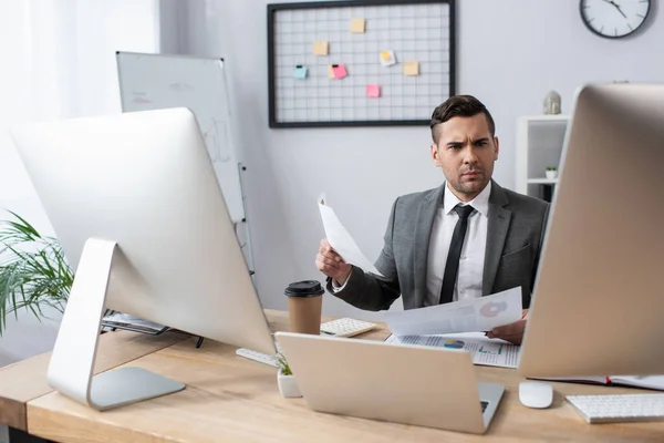 Seriöser Händler mit Papieren, während er am Arbeitsplatz in der Nähe von Laptop und Monitoren sitzt — Stockfoto