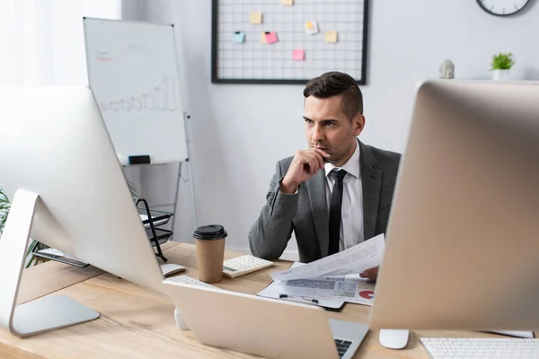 Comerciante serio sentado en el lugar de trabajo cerca de computadoras y café para ir - foto de stock