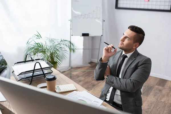 Comerciante reflexivo celebración de la pluma y mirando hacia otro lado mientras está sentado en el lugar de trabajo - foto de stock