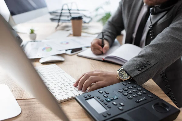 Vista recortada del comerciante hablando por teléfono fijo, escribir en el cuaderno y escribir en el ordenador portátil, borrosa primer plano - foto de stock