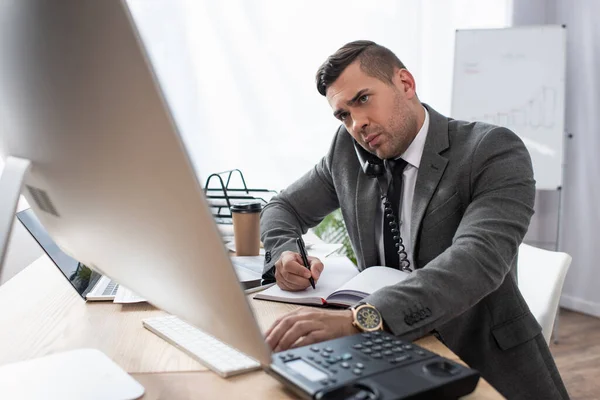 Comerciante concentrado mirando el monitor de la computadora, hablando por teléfono y la escritura en el cuaderno, borrosa primer plano - foto de stock