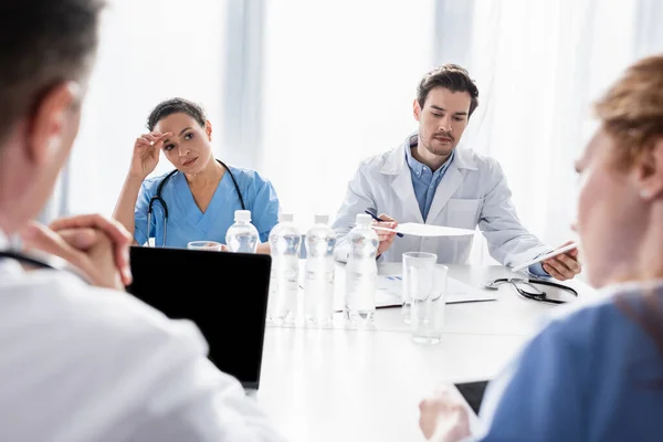 Multiethnic doctors and nurses working with papers and devices near colleagues on blurred foreground in clinic — Stock Photo