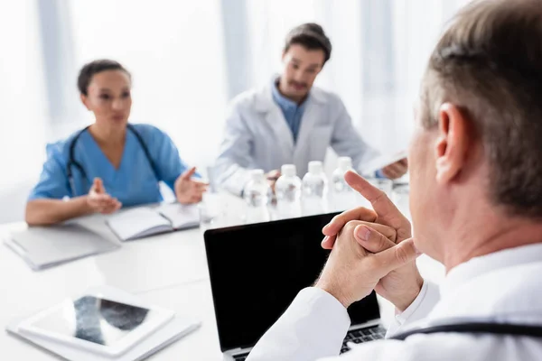Arzt zeigt mit Finger auf Laptop und multiethnische Kollegen am Tisch vor verschwommenem Hintergrund — Stockfoto