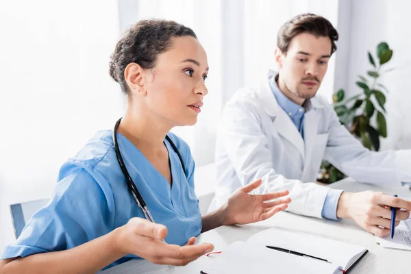 Enfermeira afro-americana olhando para longe perto de médico e notebook sobre fundo borrado — Fotografia de Stock