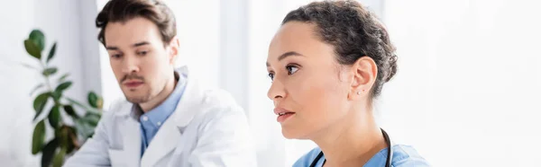Enfermera afroamericana mirando hacia otro lado cerca del médico sobre fondo borroso en la clínica, pancarta - foto de stock