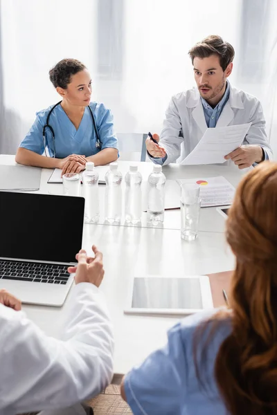Doctor holding paper and talking to multiethnic colleagues near devices and water on table — Stock Photo