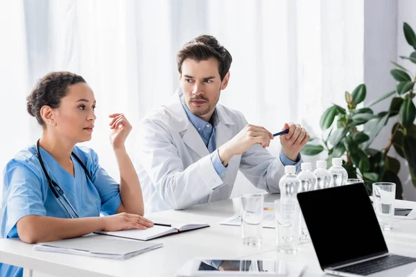 Medico guardando infermiere afro-americano vicino a documenti, dispositivi e acqua in clinica — Foto stock