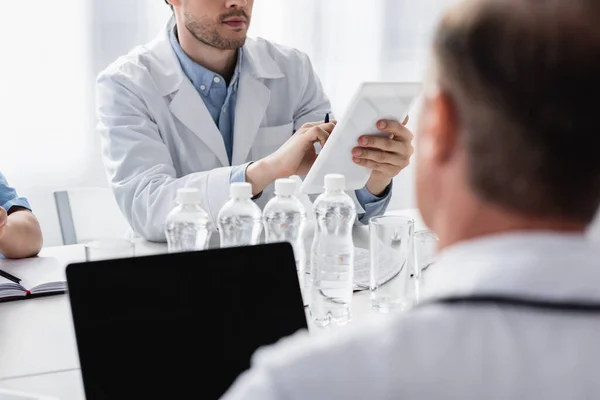 Cropped view of doctor using digital tablet near bottles of water and colleague on blurred foreground — Stock Photo