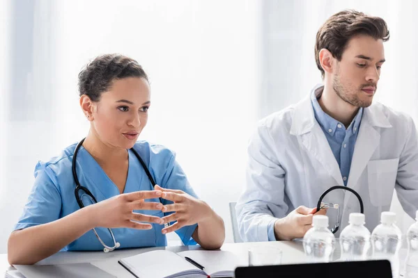 African american nurse talking near colleague with stethoscope, notebook and bottles of water on blurred foreground — Stock Photo