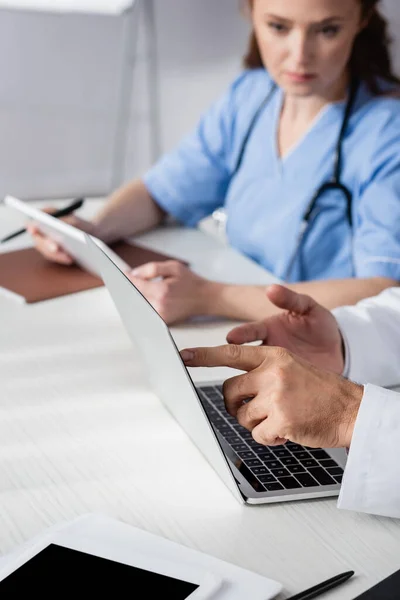 Médico apuntando a la computadora portátil cerca de la tableta digital y enfermera en fondo borroso en el hospital - foto de stock