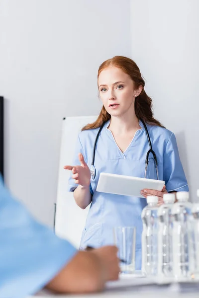 Nurse pointing with finger while holding digital tablet near colleagues and bottles of water on blurred foreground — Stock Photo