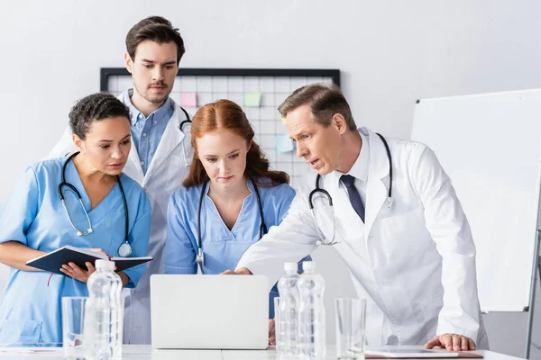 Multiethnische Ärzte und Krankenschwestern mit Laptop in Wassernähe auf verschwommenem Vordergrund — Stockfoto