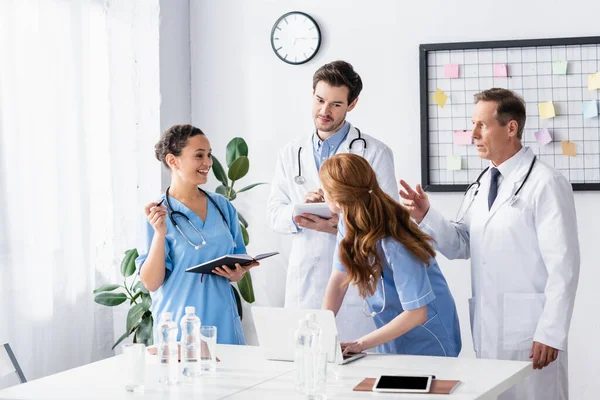 Infermiere afro-americane sorridenti con taccuino che guardano colleghi con dispositivi in clinica — Foto stock