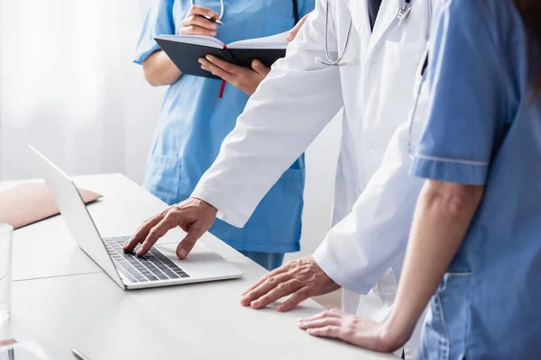 Cropped view of doctor in white coat using laptop near nurses with notebook — Stock Photo