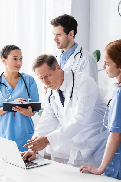 Doctor señalando con el dedo a la computadora portátil cerca de colegas multiculturales en el hospital - foto de stock