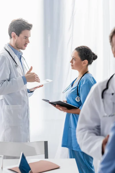 Arzt und afrikanisch-amerikanische Krankenschwester mit Notizbuch und digitalem Tablet im Gespräch mit Kollegen auf verschwommenem Vordergrund — Stockfoto