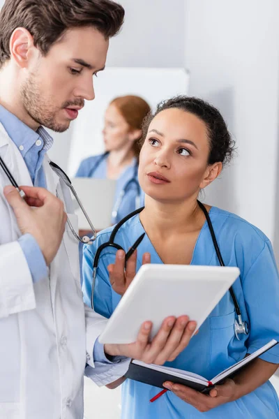 African american nurse with notebook standing near doctor with digital tablet on blurred foreground — Stock Photo