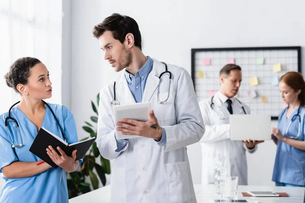 Multiethnic hospital staff with notebook and digital tablet talking near colleagues with laptop on blurred background — Stock Photo