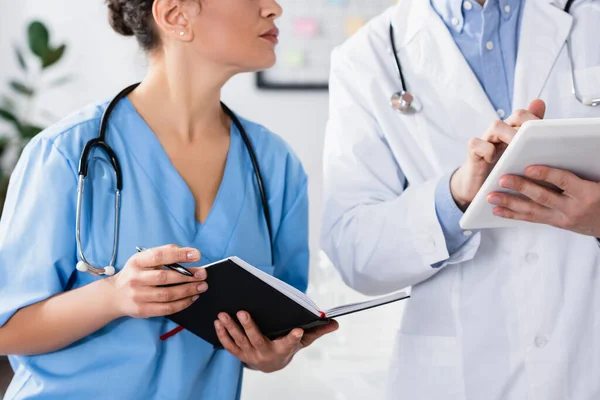 Ausgeschnittene Ansicht einer afrikanisch-amerikanischen Krankenschwester mit Stift und Notizbuch, die in der Nähe eines Arztes mit digitalem Tablet im Krankenhaus steht — Stockfoto