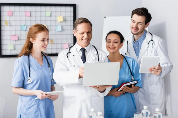 Fröhliche multiethnische Ärzte mit Geräten und Notizbuch im Krankenhaus — Stockfoto