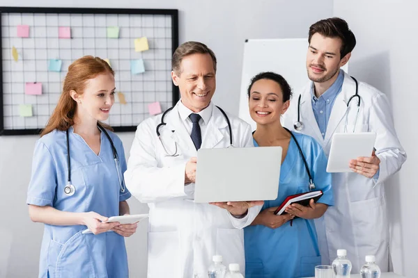 Alegre médicos multiétnicos con dispositivos y cuaderno de trabajo en el hospital — Stock Photo