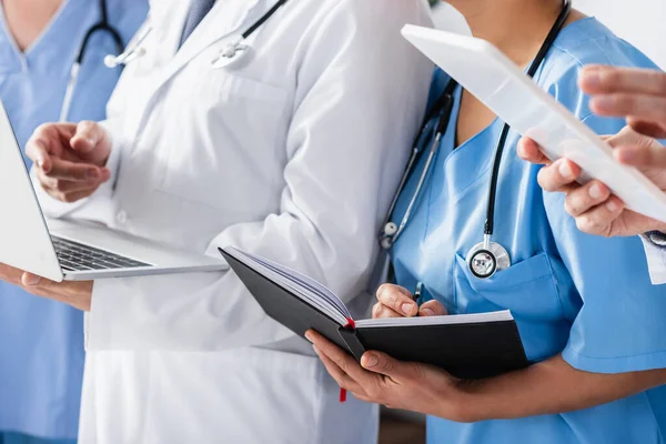 Ausgeschnittene Ansicht einer Krankenschwester, die auf einem Notizbuch in der Nähe von Kollegen schreibt, mit Geräten auf verschwommenem Hintergrund — Stockfoto