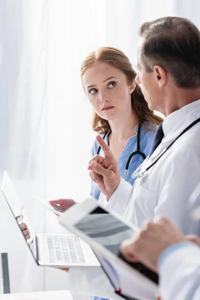 Doctor with laptop pointing with finger near nurse with digital tablet and colleague on blurred foreground — Stock Photo