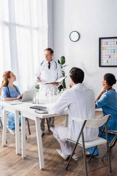 Arzt mit Papiermappe zeigt mit der Hand in die Nähe multiethnischer Kollegen und Geräte auf dem Tisch — Stockfoto