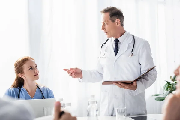 Doctor holding paper folder near nurse with laptop and colleagues on blurred foreground — Stock Photo