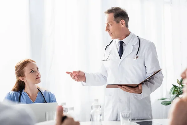 Doctor sosteniendo la carpeta de papel cerca de la enfermera con el ordenador portátil y colegas en primer plano borrosa - foto de stock