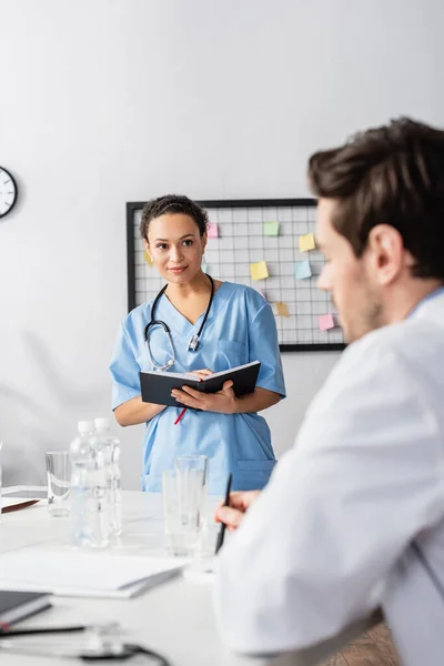 African american nurse with notebook looking at colleague near papers and water on blurred foreground — Stock Photo