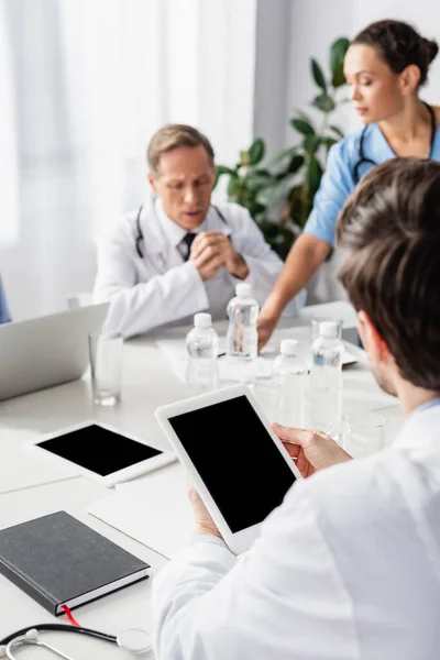 Tablette numérique avec écran blanc dans les mains du médecin près de collègues multiethniques et des appareils sur fond flou — Photo de stock