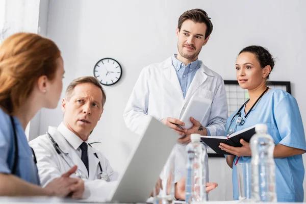 Personnel hospitalier multiethnique avec ordinateur portable et appareils travaillant près de l'eau au premier plan flou sur la table — Photo de stock