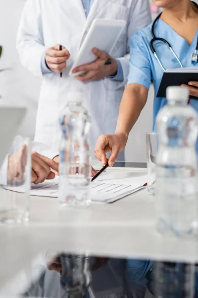Cropped view of african american nurse pointing at papers near colleagues with digital tablet and bottles of water on blurred foreground — Stock Photo