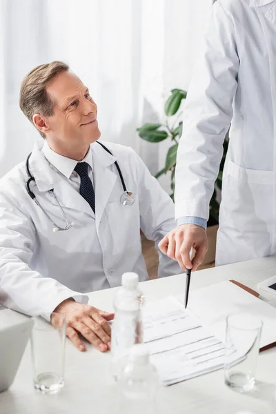 Médico sonriente sentado cerca de un colega señalando papeles y botellas de agua en un primer plano borroso - foto de stock