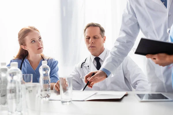 Doctor and nurse sitting near colleague pointing with hand at papers near bottles and digital tablet on blurred foreground — Stock Photo