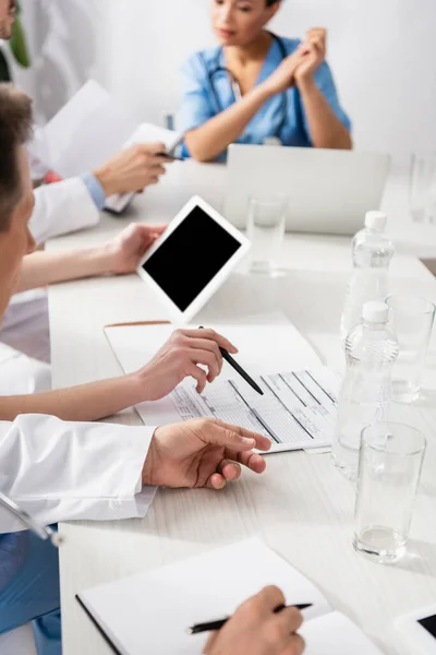 Papeles y botellas de agua en la mesa cerca del personal del hospital multiétnico con cuadernos y dispositivos sobre fondo borroso - foto de stock