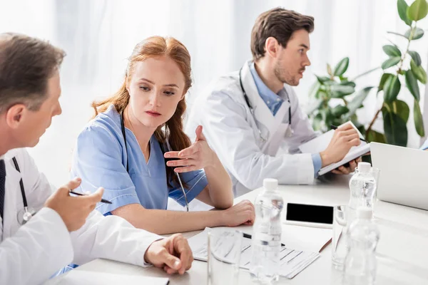 Krankenschwester zeigt mit der Hand in die Nähe des Arztes, Papiere und Wasserflaschen im verschwommenen Vordergrund — Stockfoto