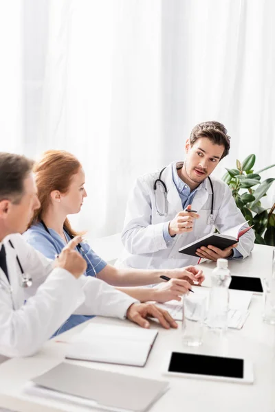 Médico segurando caneta e notebook perto de colegas, dispositivos e papéis em primeiro plano desfocado — Fotografia de Stock