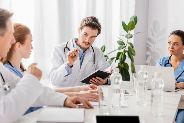 Doctor with notebook looking at colleagues near african american nurse with laptop in clinic — Stock Photo