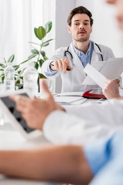 Doctor with papers pointing with finger near colleagues with digital tablet on blurred foreground — Stock Photo