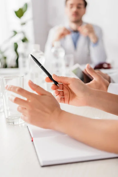 Doctor holding pen near notebook and colleagues on blurred foreground at table — Stock Photo
