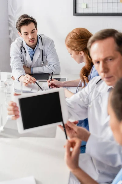 Arzt zeigt mit digitalem Tablet im unscharfen Vordergrund auf Papiere in der Nähe von Krankenschwester und Kollegen — Stockfoto