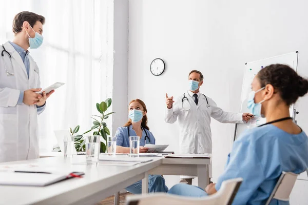 Médico em máscara médica apontando com o dedo perto de colegas multiétnicos com papéis e tablet digital na clínica — Fotografia de Stock