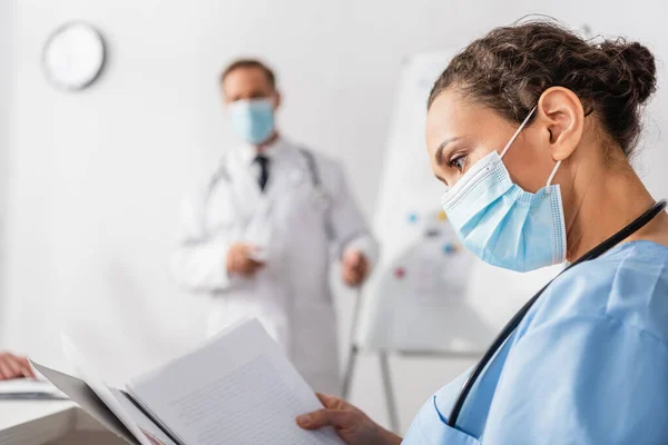 Enfermeira afro-americana em máscara médica olhando para papéis na pasta perto colega em fundo turvo — Fotografia de Stock