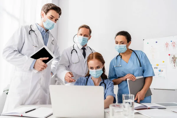 Médicos e enfermeiros multiétnicos em máscaras médicas usando laptop enquanto trabalham perto de papéis em primeiro plano desfocado — Fotografia de Stock